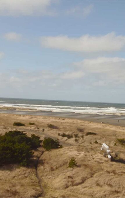 The image shows a coastal scene with grassy sand dunes in the foreground, leading to a beach and the ocean in the background. A building is located on the left side, and a road curves towards the beach on the right. The sky is partly cloudy.