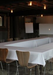 A room with a U-shaped arrangement of tables covered with white tablecloths. Surrounding the tables are wooden chairs. The room features exposed beams on the ceiling and several hanging light bulbs. There are whiteboards and a flipchart at the far end of the room.