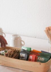 A hand reaches for sugar packets in a wicker basket next to an assortment of tea bags on a white table. Three white mugs and a jar filled with stir sticks are also on the table, beside a coffee urn.