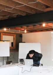 A person is bending over a laptop on a table in a room set up for a meeting or conference. There are projection screens, flip charts, several glasses, and a bottle of water on the tables. The ceiling has exposed beams and hanging light bulbs.