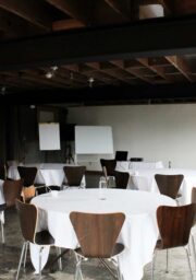 A meeting room with round tables covered in white tablecloths, surrounded by wooden chairs. In the background, there are whiteboards and flip charts. The room has a rustic feel with exposed beams and large windows that bring in natural light.