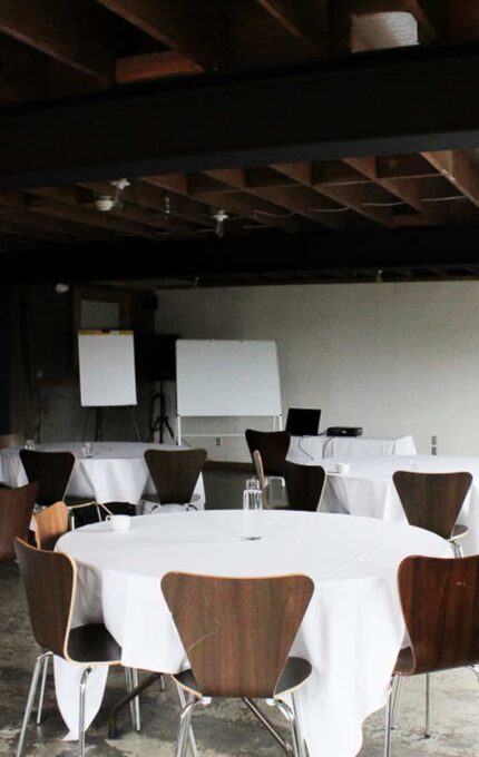 A meeting room with round tables covered in white tablecloths, surrounded by wooden chairs. In the background, there are whiteboards and flip charts. The room has a rustic feel with exposed beams and large windows that bring in natural light.