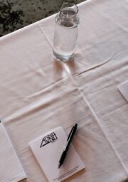 A table setup with a white tablecloth features three pads of paper each with a black pen placed on top. There are three glasses of water, one next to each pad.