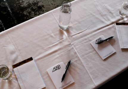 A table setup with a white tablecloth features three pads of paper each with a black pen placed on top. There are three glasses of water, one next to each pad.