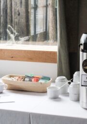 A beverage station with a coffee dispenser labeled "Forest Dark," packets of tea, sugar, and plastic stirrers on a table covered with a white tablecloth. Four white mugs and two small containers are also on the table, situated near a window with a curtain.