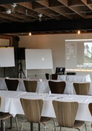 A conference room featuring rows of wooden chairs and long tables covered with white tablecloths. The room has a ceiling with exposed beams. At the front, there is a projector displaying an image, along with whiteboards and flip charts.