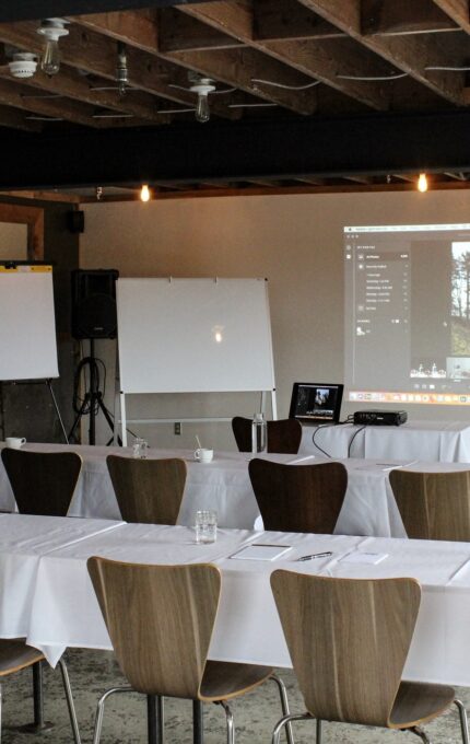 A conference room featuring rows of wooden chairs and long tables covered with white tablecloths. The room has a ceiling with exposed beams. At the front, there is a projector displaying an image, along with whiteboards and flip charts.