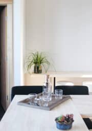 A minimalist dining area features a light wooden table with a wooden tray holding a glass pitcher and four glasses. In the background, there is a green potted plant on a shelf, and a small pot of succulents sits on the table.