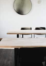 A minimalistic room with two wooden tables and black chairs arranged in two rows. A round mirror with a gradient design hangs on the white wall above the furniture. The flooring is dark wood.