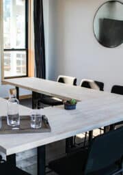 A modern office meeting room with a U-shaped wooden table, black chairs, and a round mirror on the wall. A bottle, glasses, and a small plant are placed on a wooden tray on the table. There is a large window with natural light.