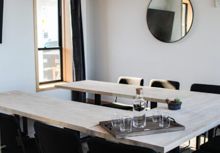 A modern conference room with light wooden rectangular tables arranged in a U-shape. Empty glasses and a carafe of water sit on a tray in the center. Dark chairs surround the tables, and a round mirror hangs on the wall. A window lets in natural light.