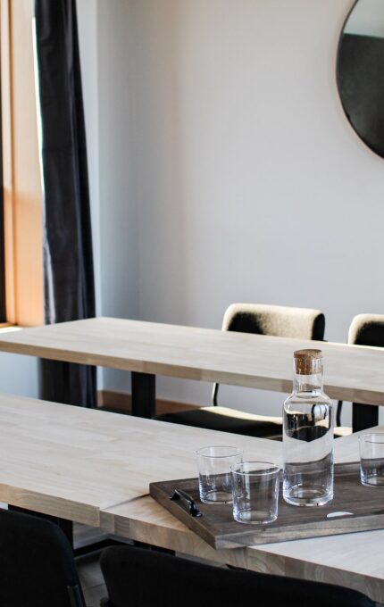 A modern conference room with light wooden rectangular tables arranged in a U-shape. Empty glasses and a carafe of water sit on a tray in the center. Dark chairs surround the tables, and a round mirror hangs on the wall. A window lets in natural light.