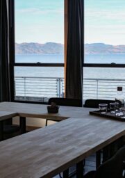 A conference room with a wooden U-shaped table and black chairs. The room has large windows offering a view of a body of water and distant mountains. A tray with a carafe, glasses, and a few small items is on the table.