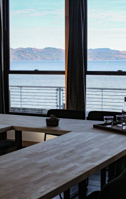 A conference room with a wooden U-shaped table and black chairs. The room has large windows offering a view of a body of water and distant mountains. A tray with a carafe, glasses, and a few small items is on the table.