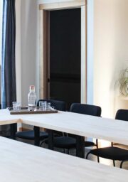 A well-lit conference room with a U-shaped arrangement of light wooden tables surrounded by black chairs. On the tables are a tray with a water jug and glasses, and a small succulent plant. A taller plant sits on a counter against the wall.