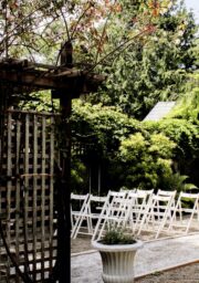 A garden with white folding chairs is set up in rows, facing a wooden arbor adorned with lattice and sparse climbing plants. The setting is surrounded by trees and greenery, with a planter positioned in front of the chairs.