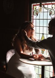 A woman in a white dress and a man in a gray shirt sit at a table in front of a stained glass window. The man gently touches the woman's face while they both smile. They are holding hands across the table and appear to be in a dimly lit setting.