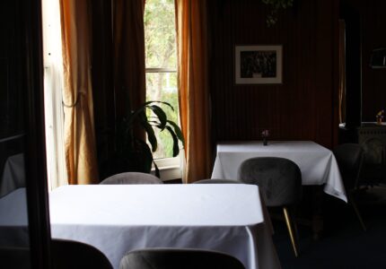 A dimly lit restaurant interior with tables covered in white cloths. Gray upholstered chairs are positioned around the tables. A window with long, golden curtains is in the background, with sunlight filtering through. A potted plant and framed picture are also visible.