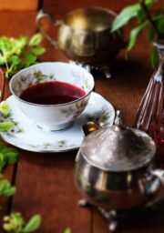 A wooden table set with a delicate floral-patterned teacup filled with red liquid, a matching saucer, and a silver sugar bowl. Surrounding them are green leaves, a silver teapot, and decorative glass vases with plants and flowers.