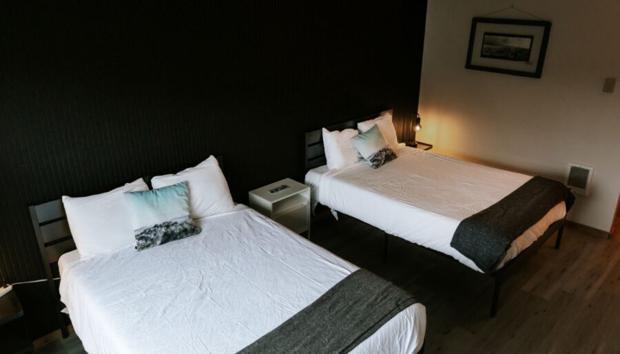 A hotel room with two neatly made double beds, each with white bedding, gray throws, and decorative pillows. There is a nightstand with a lamp between the beds and a framed picture on the white wall.