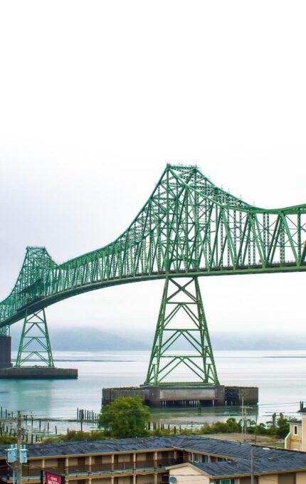 A green steel truss bridge spans across a body of water on a foggy day, connecting to land on either side. In the foreground, there are several buildings, including a beige multi-story structure. Hills can be seen in the distant background.