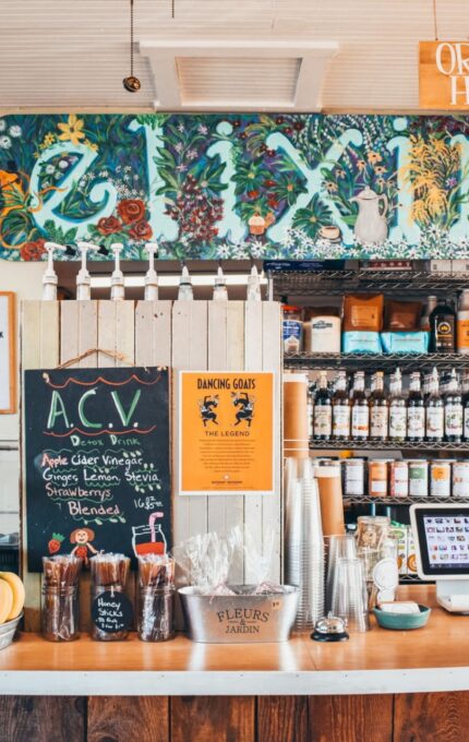 A cozy cafe counter with a variety of items displayed. Shelves behind the counter hold jars and bottles. There's a sign above the counter reading "Elixir" and a "Specials" board on the right. The counter has snacks, a fruit basket, and an order sign.