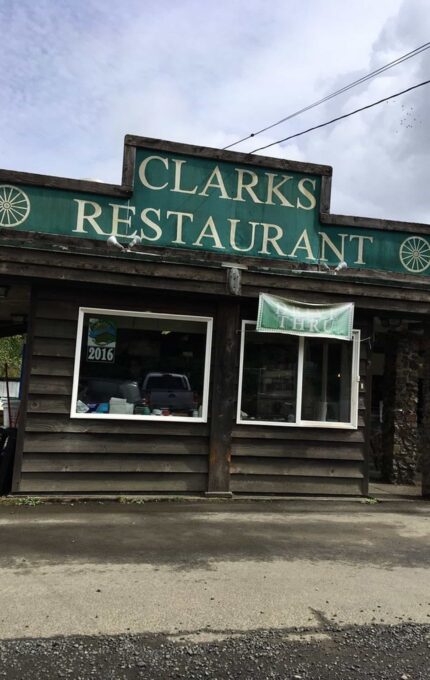 A rustic wood and stone building with a green sign reading "Clarks Restaurant" on top. The building has large windows and a 2016 award in one of the windows. A banner on the window says "THIRD." The sky is overcast, and the ground is wet.