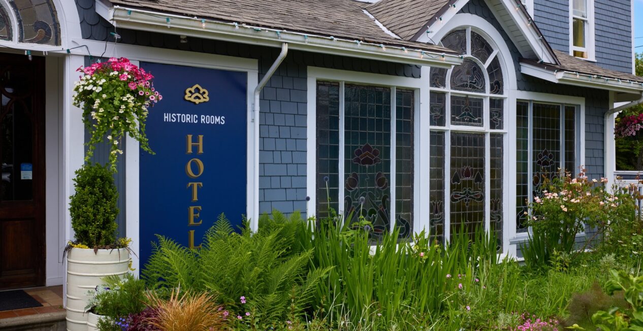 A building with blue-gray siding and white trim, featuring a sign that reads "HISTORIC ROOMS HOTEL." The entrance is adorned with potted plants and there is a landscaped garden with various green plants and flowers in the foreground.