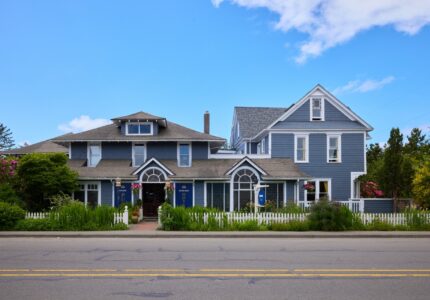 A street view of a large, two-story blue house with a white picket fence. The house has multiple gables, several windows, and two entryways with decorative arches. The yard is landscaped with green plants and flowers. The sky is clear and blue.