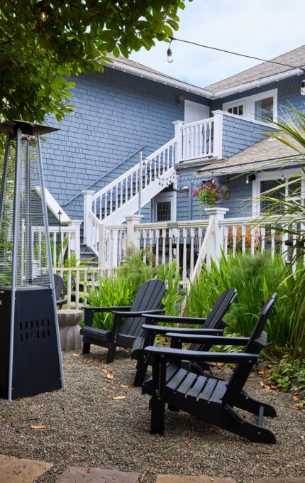 A backyard area with a gravel patio featuring black Adirondack chairs surrounding a tall propane patio heater. The yard is landscaped with various plants and trees, and a two-story gray house with white trim is in the background.