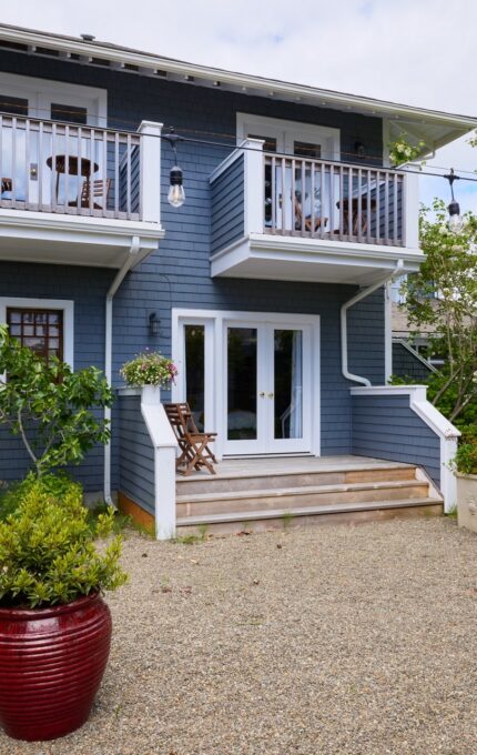 Image of a two-story house with a blue exterior and two balconies on the upper floor. The ground floor has two sets of French doors leading to a gravel patio. The patio is decorated with a large red planter and string lights. Trees and outdoor furniture are visible.