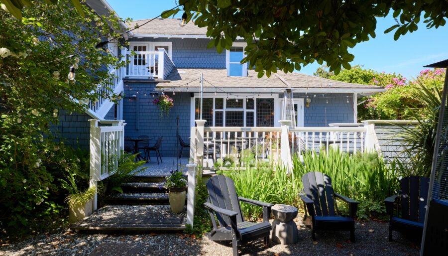 A two-story gray house features a spacious backyard deck with white railings. The deck overlooks a gravel patio area adorned with black Adirondack chairs and surrounded by lush green plants. String lights add a decorative touch under the clear blue sky.