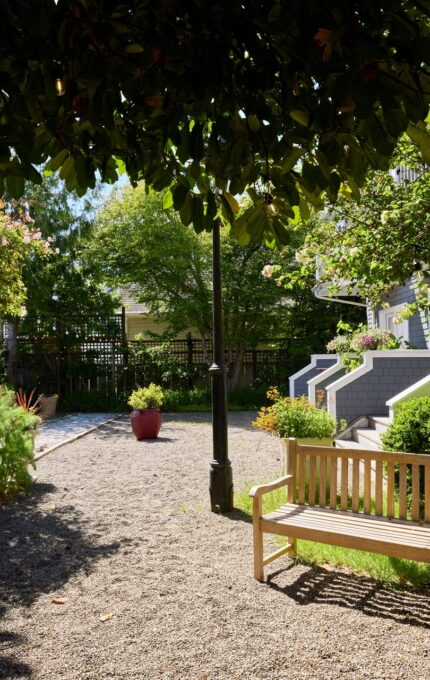 A sunny garden scene with a wooden bench on a gravel path. To the left, there is a wooden arbor with climbing plants. In the background, there are stairs leading up to a light green house partially visible through trees and shrubs.