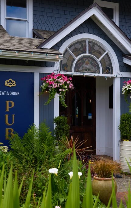 A blue building with signs for a pub and hotel. The entrance has a wooden door, hanging flower baskets, and large potted plants. The roof has grey shingles, and there's decorative trim around the door. "Eat & Drink" and "Hotel Rooms" signs are visible.