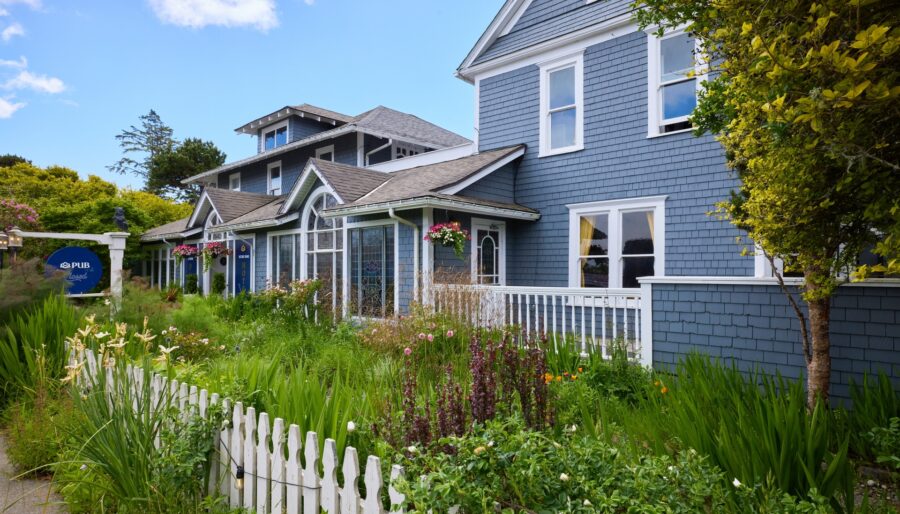 A blue two-story house with a white picket fence and a lush garden in front. The house features large windows, a covered porch, and second-story balconies. Flowering plants and greenery fill the garden area. Blue sky with scattered clouds is in the background.