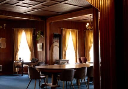 A warmly lit room with wooden paneling, a high ceiling, and blue carpet. Furnished with various tables and chairs, the space features large windows with sheer curtains allowing streams of natural light. A fireplace is visible on the far side of the room.