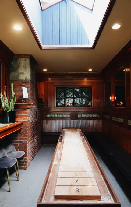 A room with wooden walls and a skylight ceiling featuring a shuffleboard table in the center. A potted plant sits on a brick structure to the left. The room has decorative stained glass windows and various lit wall lamps. Two cushioned chairs are in view.