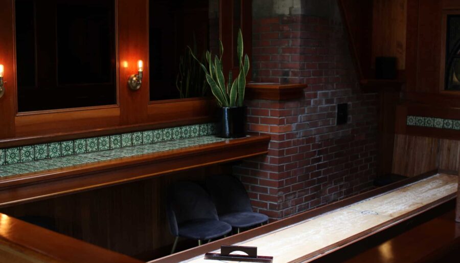 A room with a shuffleboard table, brick and wood paneling, green tiled trim, and two black chairs. There is a potted plant on a shelf along the wall, which features dark wooden panels and two wall sconces with warm lighting.