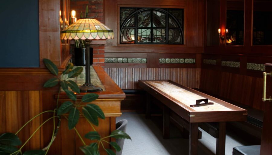 A small room with wood-paneled walls and a long wooden table. The room features stained glass windows and a stained glass lamp on a wooden ledge. There is a small potted plant on the left and a shuffleboard game on the table.