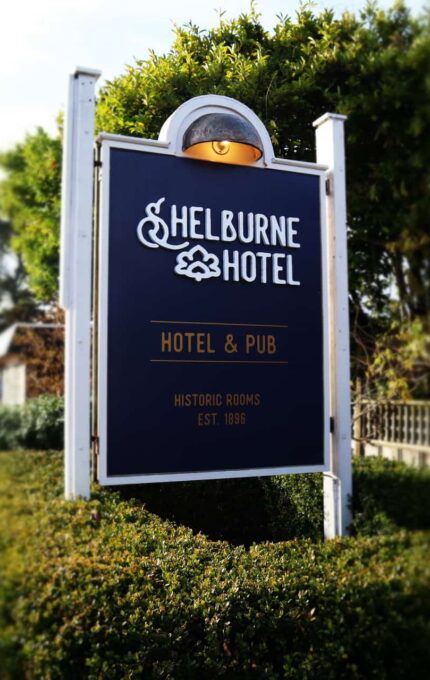 Signboard for Shelburne Hotel stands amidst greenery. It mentions "Hotel & Pub" and "Historic Rooms Est. 1896". The area around it shows parked cars and a pathway beside a fence.