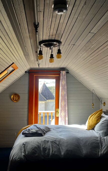 A cozy attic bedroom with a sloped wooden ceiling, a double bed with white linens and yellow pillows, a wall-mounted light fixture, a small side table, and a fan. A door at the end of the room leads to a balcony, and a window is set into the ceiling.