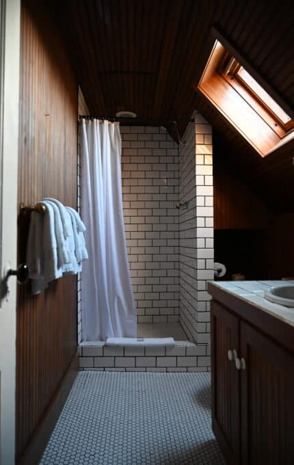 The image shows an attic bathroom with wooden walls and a ceiling. There is a white-tiled shower area with a white curtain and an overhead shower. A skylight window lets in natural light. The floor has small hexagonal white tiles, and there's a folded towel on the wall.