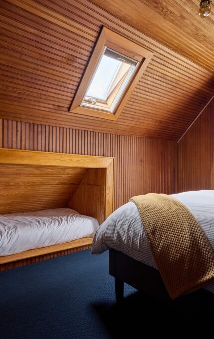 A cozy attic bedroom with wooden paneling features a single bed built into the wall on the left and a double bed with white linens and a yellow blanket on the right. The room has a skylight window and a stained glass window offering ample natural light.