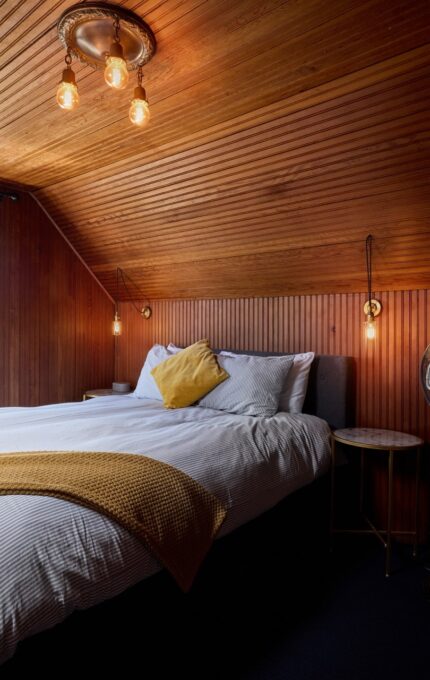 A cozy bedroom with wooden paneled walls and ceiling features a neatly made bed with white linens, a yellow pillow, and a yellow throw. There is a colorful stained glass window, a round table with a lamp, a fan, and a blue upholstered chair.