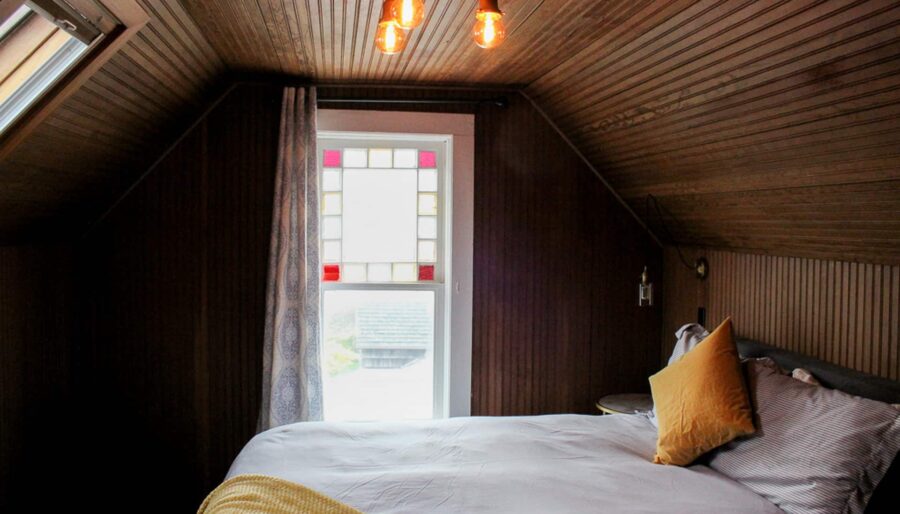 A cozy attic bedroom with wood-paneled walls and ceiling. The room features a bed with white linens, a yellow pillow, a small side table, and a hanging light fixture with exposed bulbs. The window has decorative glass panes and the room is lit naturally from the skylight.