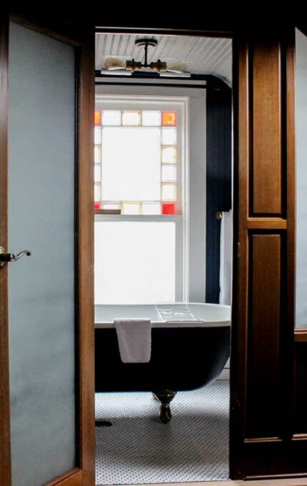 A bathroom under a sloped ceiling features a wooden door partially open, revealing a clawfoot bathtub. Natural light enters through a window with colorful stained glass panels. A skylight in the ceiling adds additional light to the room.