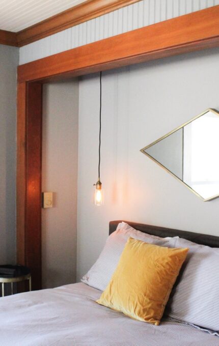 A bedroom with a bed against a gray wall, adorned with a yellow pillow on top of gray bedding. Above the bed is a rectangular mirror. Two pendant lights with exposed bulbs hang on either side of the bed. A small round side table is placed beside the bed.