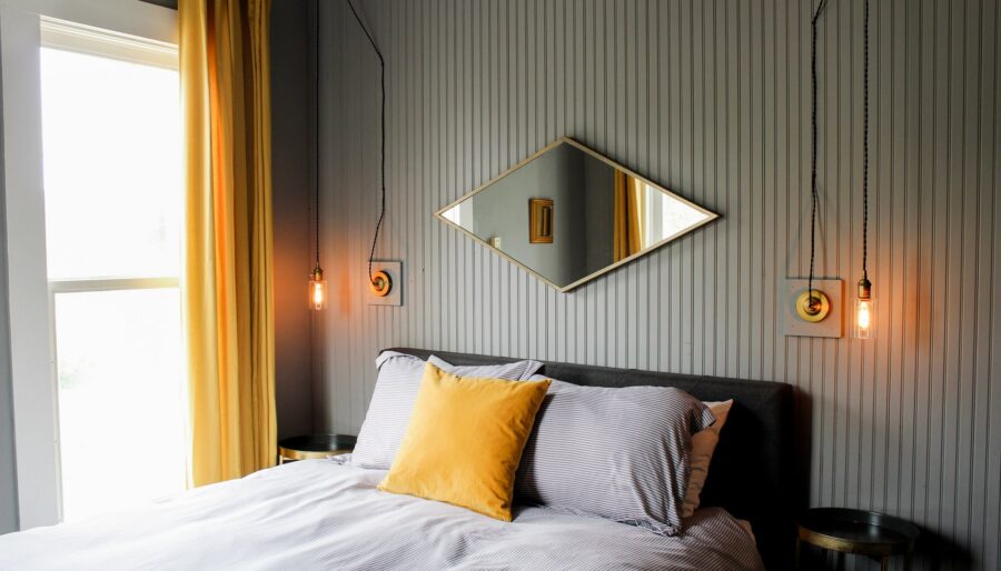 A bedroom features a bed with a gray headboard, striped bedding, and a yellow pillow. Above the bed is a diamond-shaped mirror with hanging pendant lights on either side. Yellow curtains frame a large window. The walls are gray with vertical paneling.