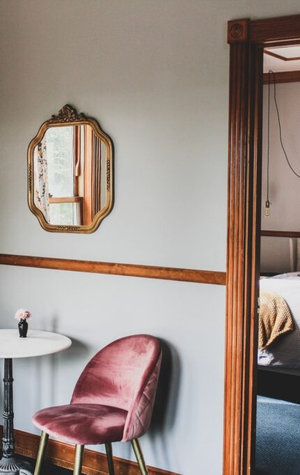 A cozy sitting area with two pink velvet chairs and a small round table is in the foreground. A gold-framed mirror hangs on the wall. An open doorway leads to a bedroom with a large bed, decorated with pillows and a throw blanket.