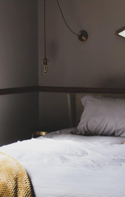 A bedroom with a neatly made bed featuring gray and white striped bedding, a yellow pillow, and a yellow blanket draped over the edge. There's a window with floral curtains, a wall-mounted light fixture, and a hexagonal mirror on the wall.